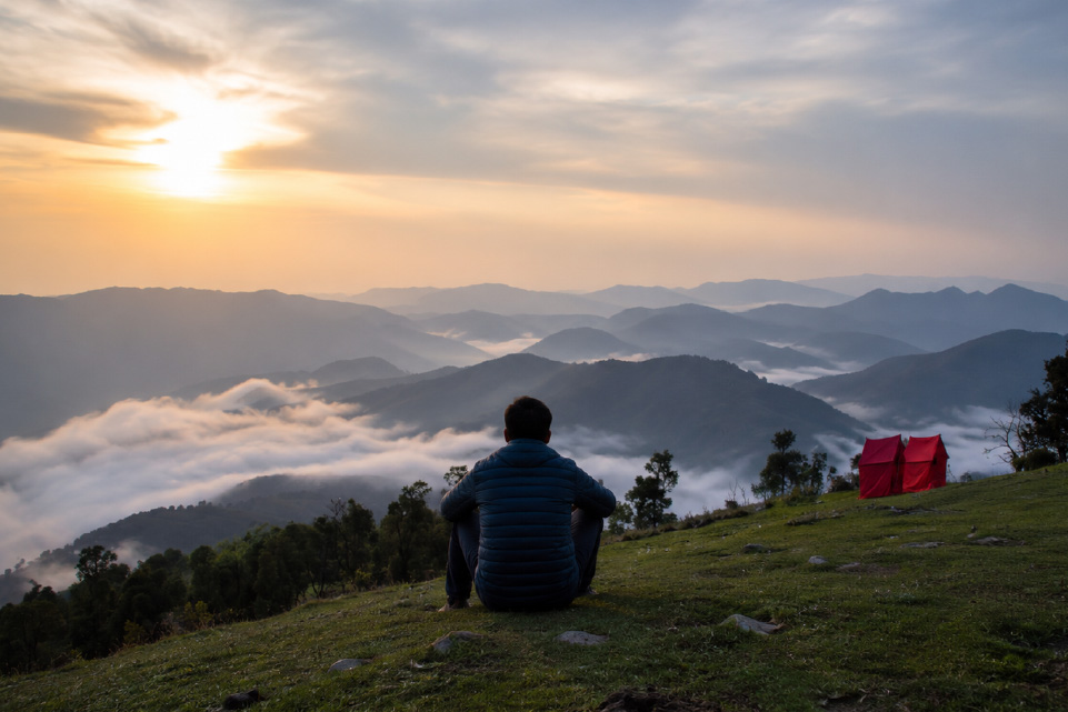 Trekker enjoying a sunrise view from Nag Tibba with mist-covered Himalayan mountains and camping tents in Uttarakhand