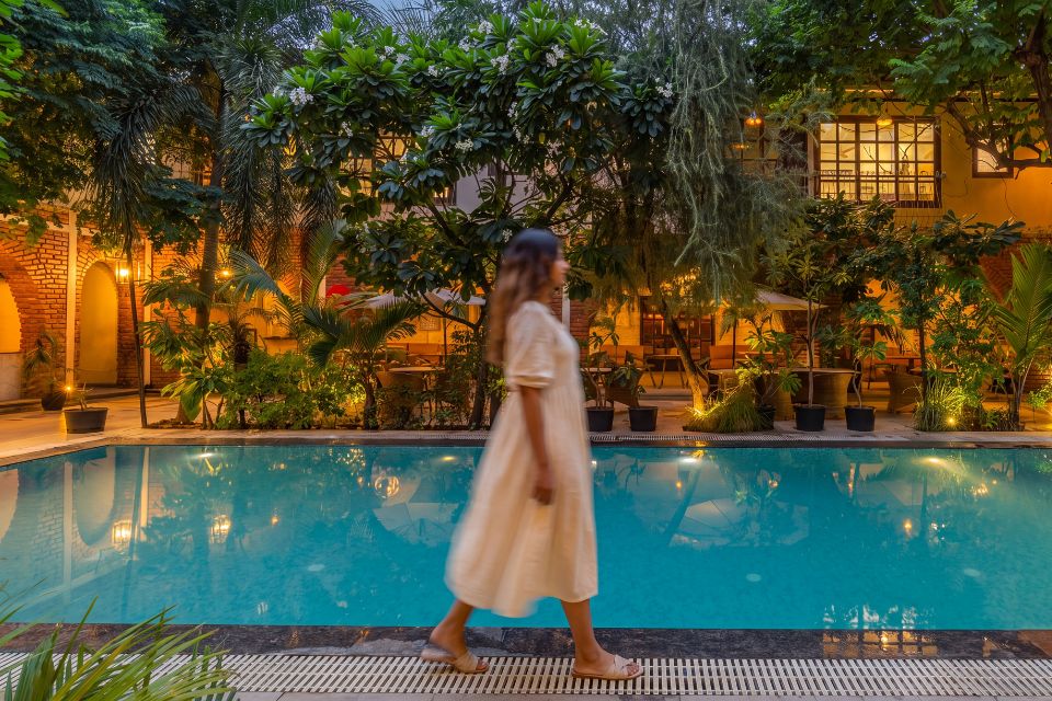 Woman in a white dress walks past the lit courtyard pool at The Hosteller BAM Udaipur at dusk, with trees and brick arches behind